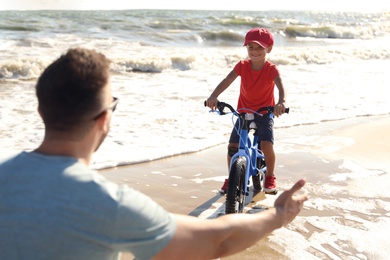 Photo of Happy father teaching son to ride bicycle on sandy beach near sea