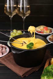 Photo of Dipping different products into fondue pot with melted cheese on black wooden table