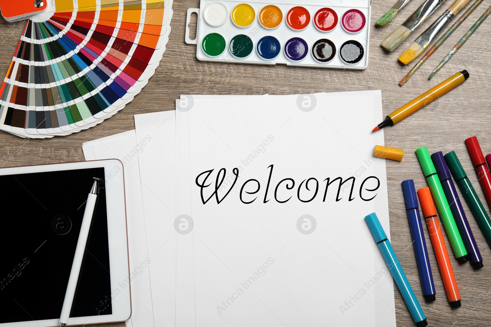 Image of Paper with word Welcome, colorful markers, watercolor paints, palette and tablet on wooden table, flat lay