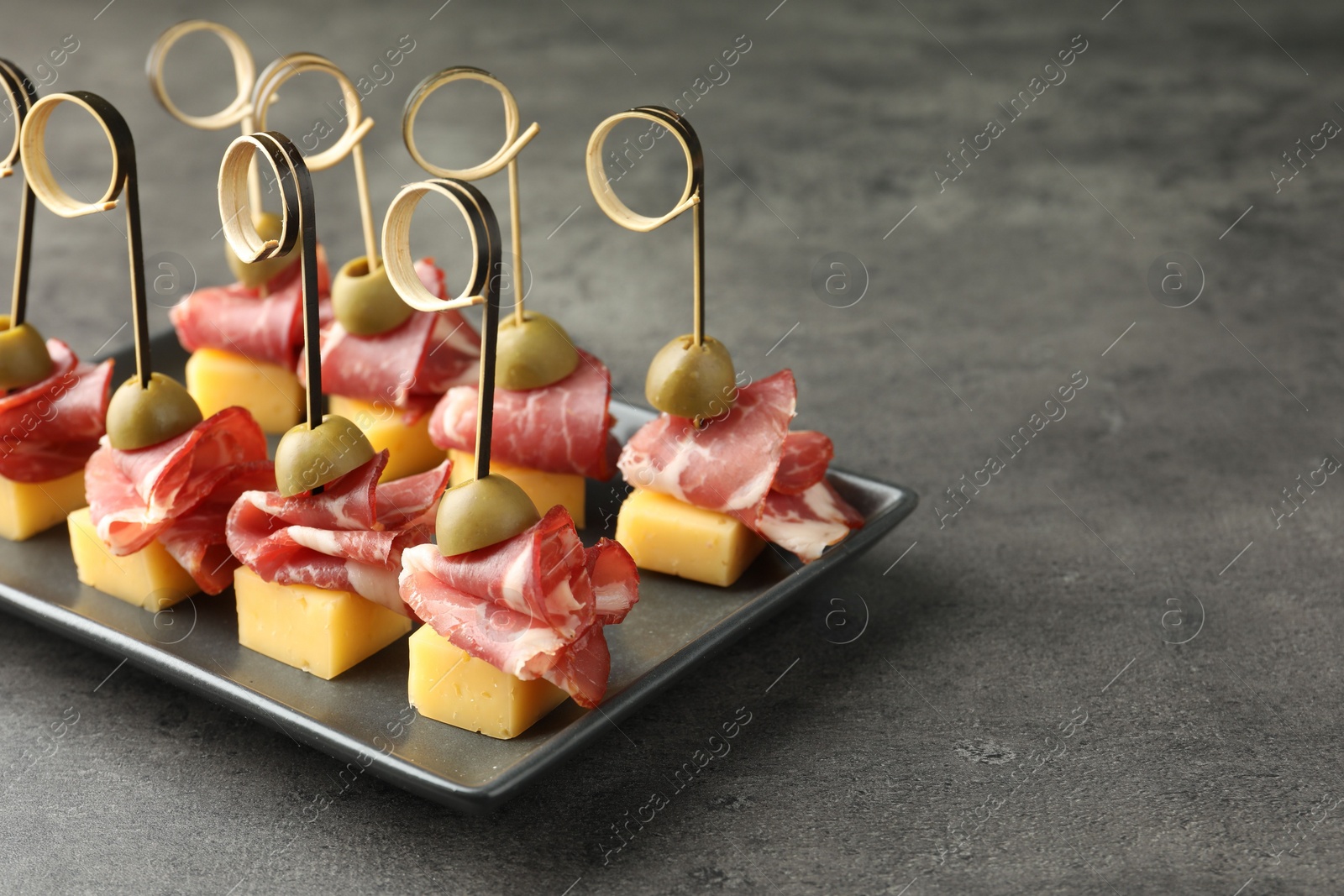 Photo of Tasty canapes with olives, prosciutto and cheese on grey table, closeup. Space for text