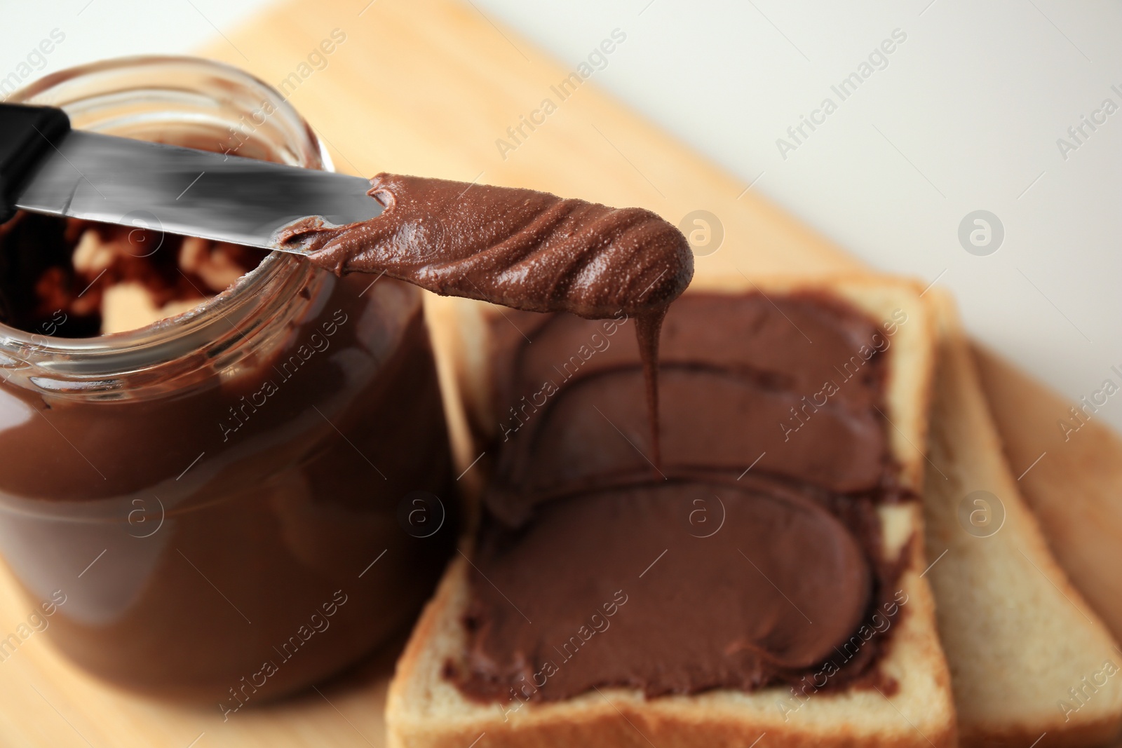 Photo of Tasty toast with chocolate paste, knife and jar on wooden board, closeup. Space for text