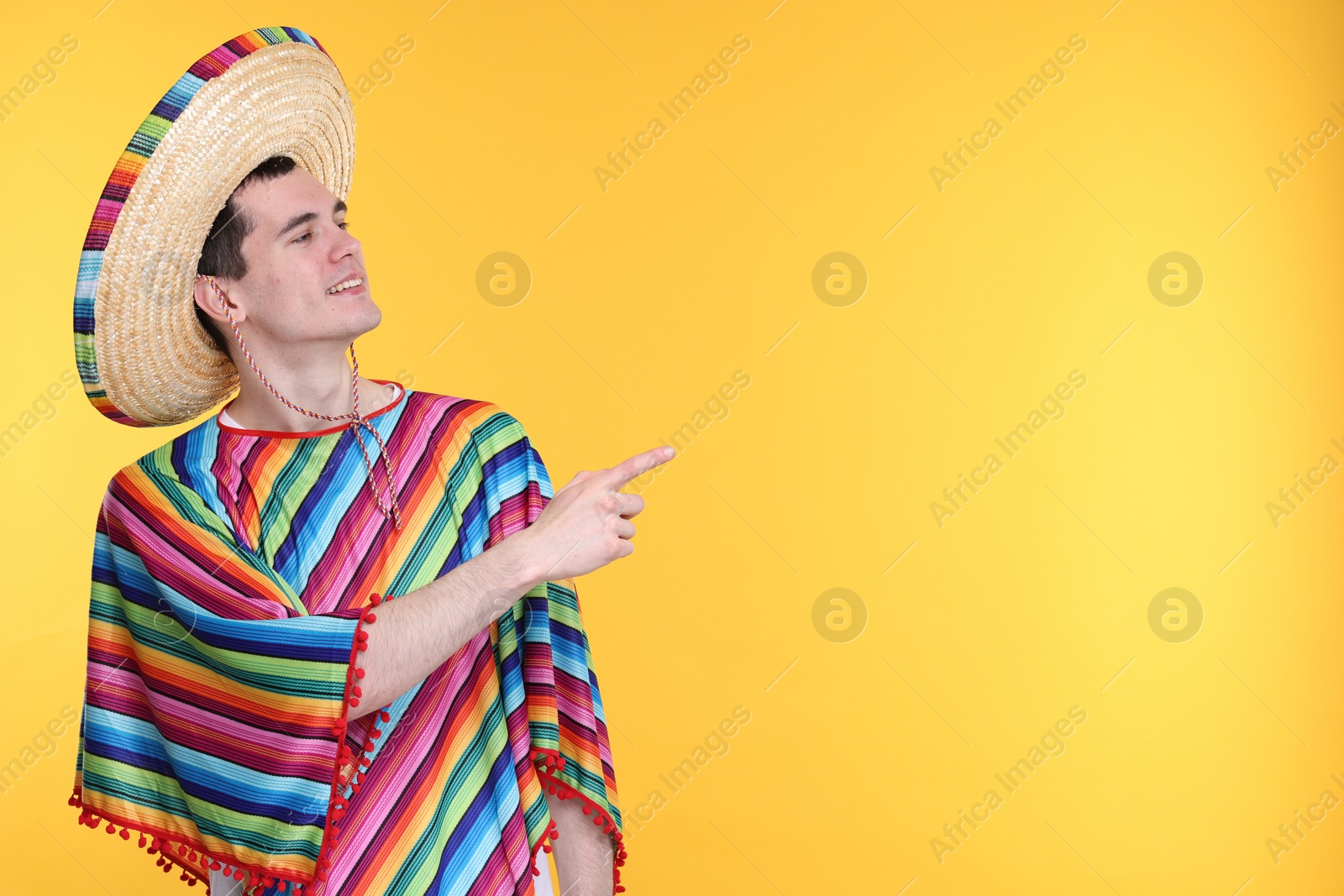 Photo of Young man in Mexican sombrero hat and poncho pointing at something on yellow background. Space for text