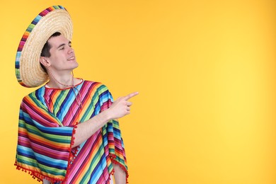 Young man in Mexican sombrero hat and poncho pointing at something on yellow background. Space for text