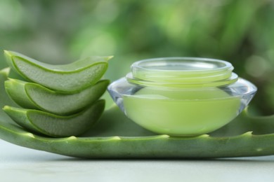 Jar with cream and cut aloe leaf on white table against blurred green background, closeup