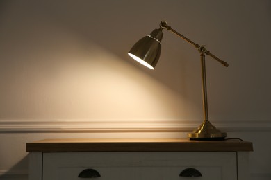 Photo of Wooden chest of drawers with modern lamp near beige wall, space for text. Interior element