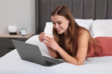 Happy woman with cup of drink and laptop on bed in bedroom