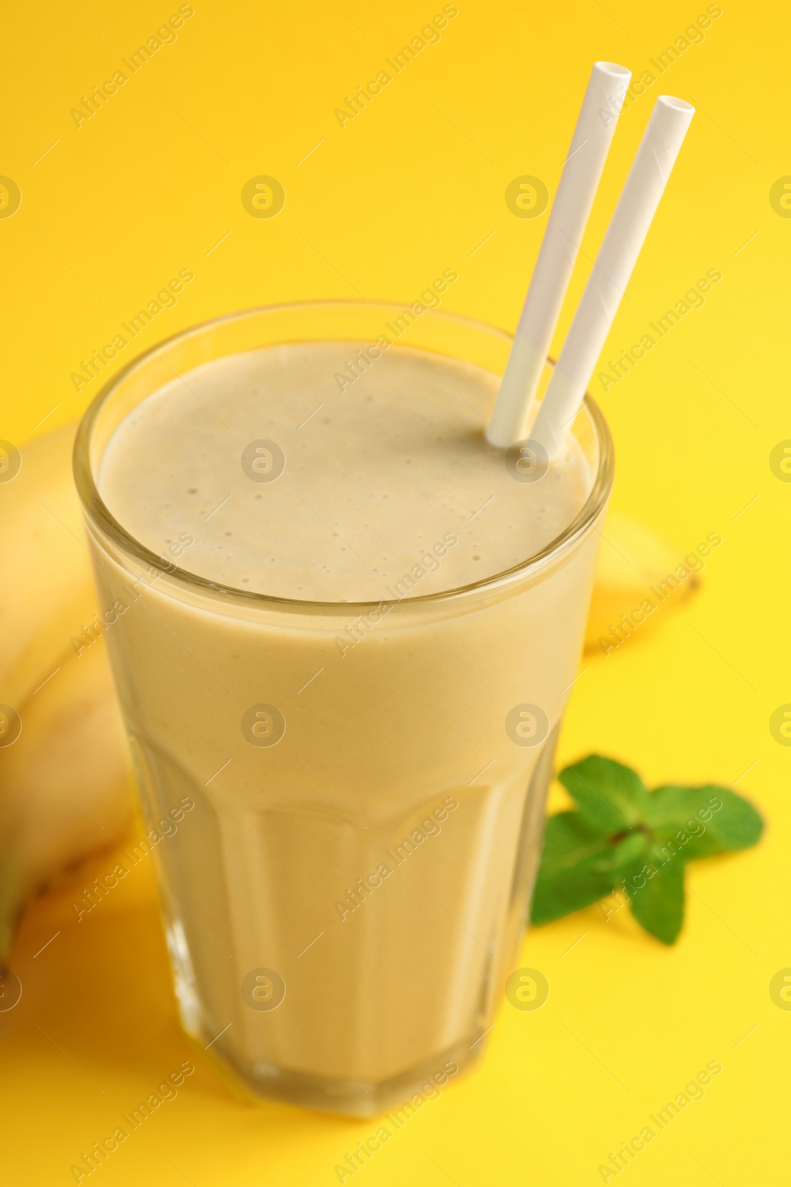 Photo of Glass of tasty banana smoothie with straws, fresh fruits and mint on yellow background, closeup