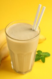 Photo of Glass of tasty banana smoothie with straws, fresh fruits and mint on yellow background, closeup