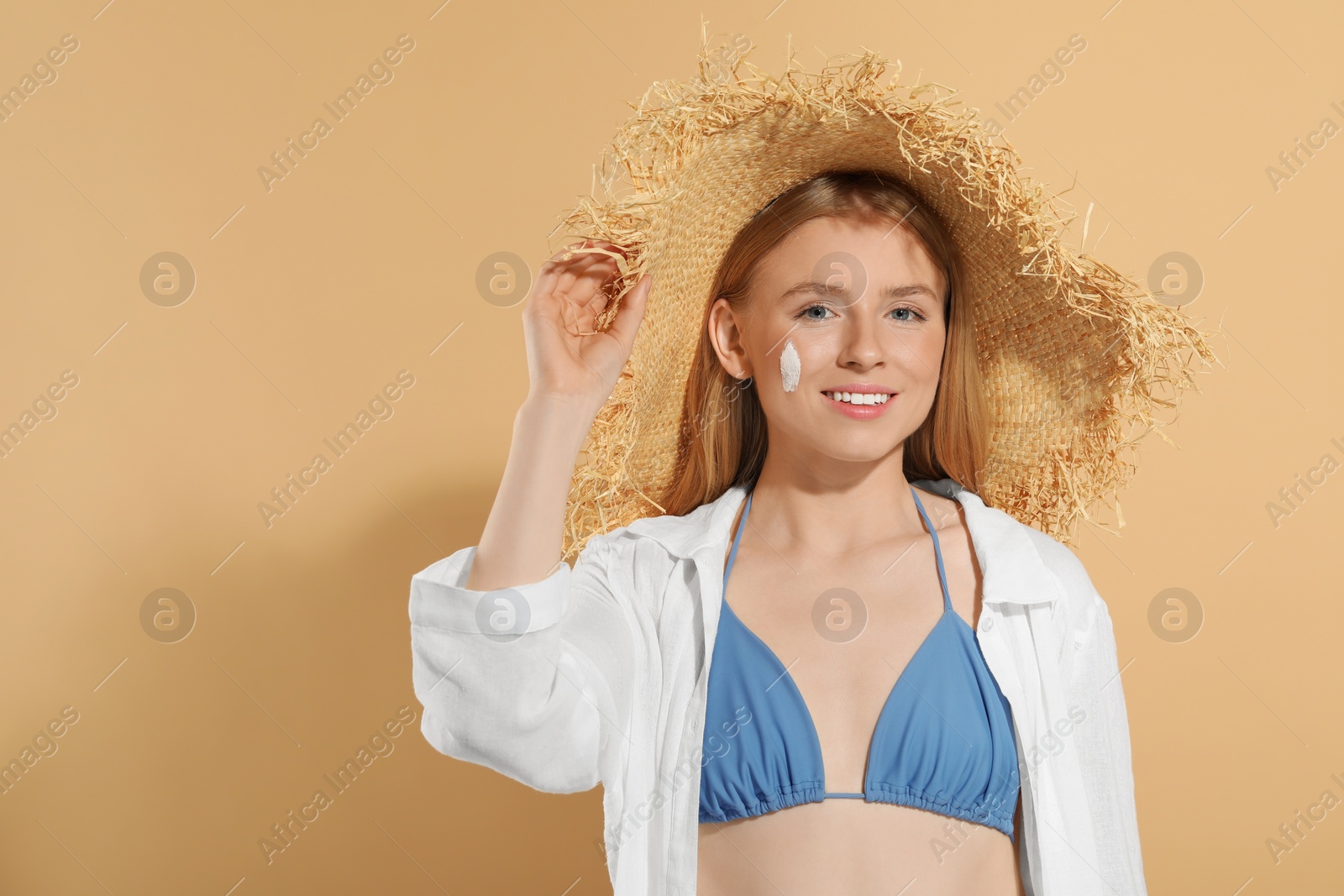 Photo of Beautiful young woman in straw hat with sun protection cream on her face against beige background, space for text