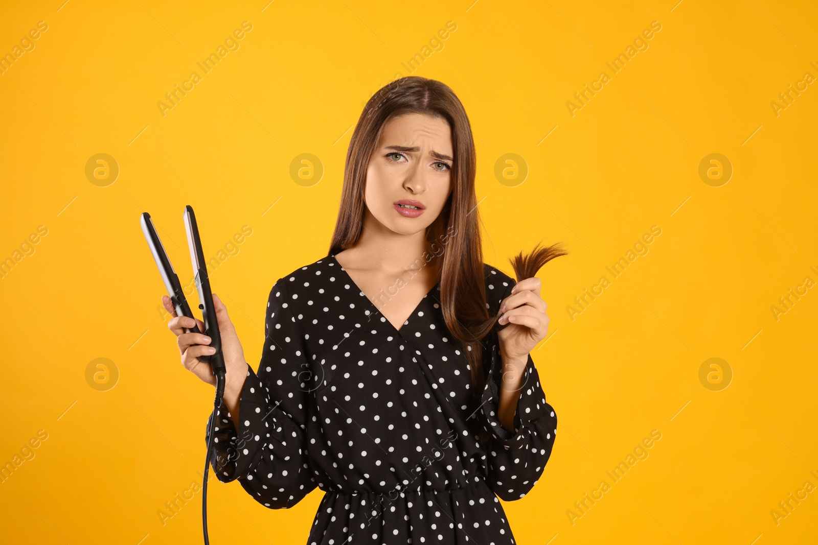 Photo of Upset young woman with flattening iron on yellow background. Hair damage