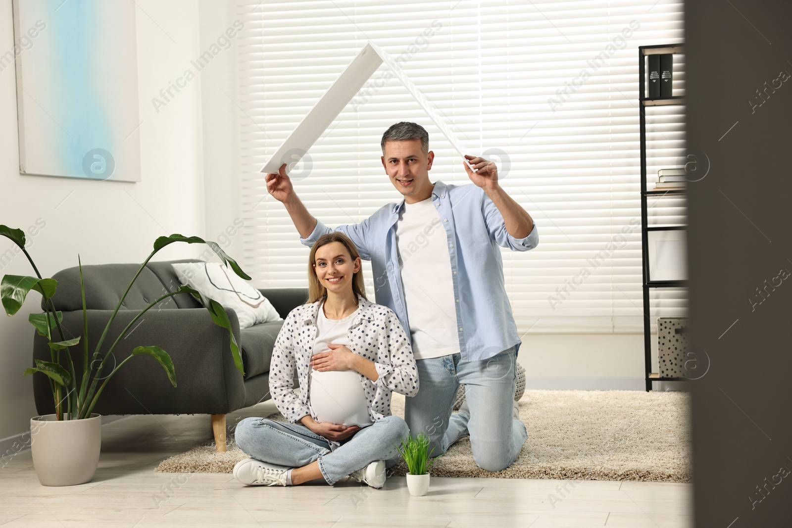 Photo of Young family housing concept. Man holding cardboard roof over his pregnant wife head at home