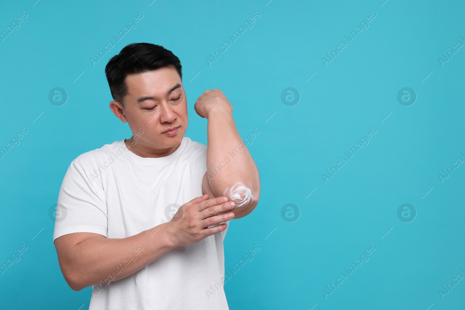 Photo of Handsome man applying body cream onto his elbow on light blue background. Space for text