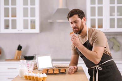 Photo of Man with freshly baked cookies watching online cooking course via tablet in kitchen