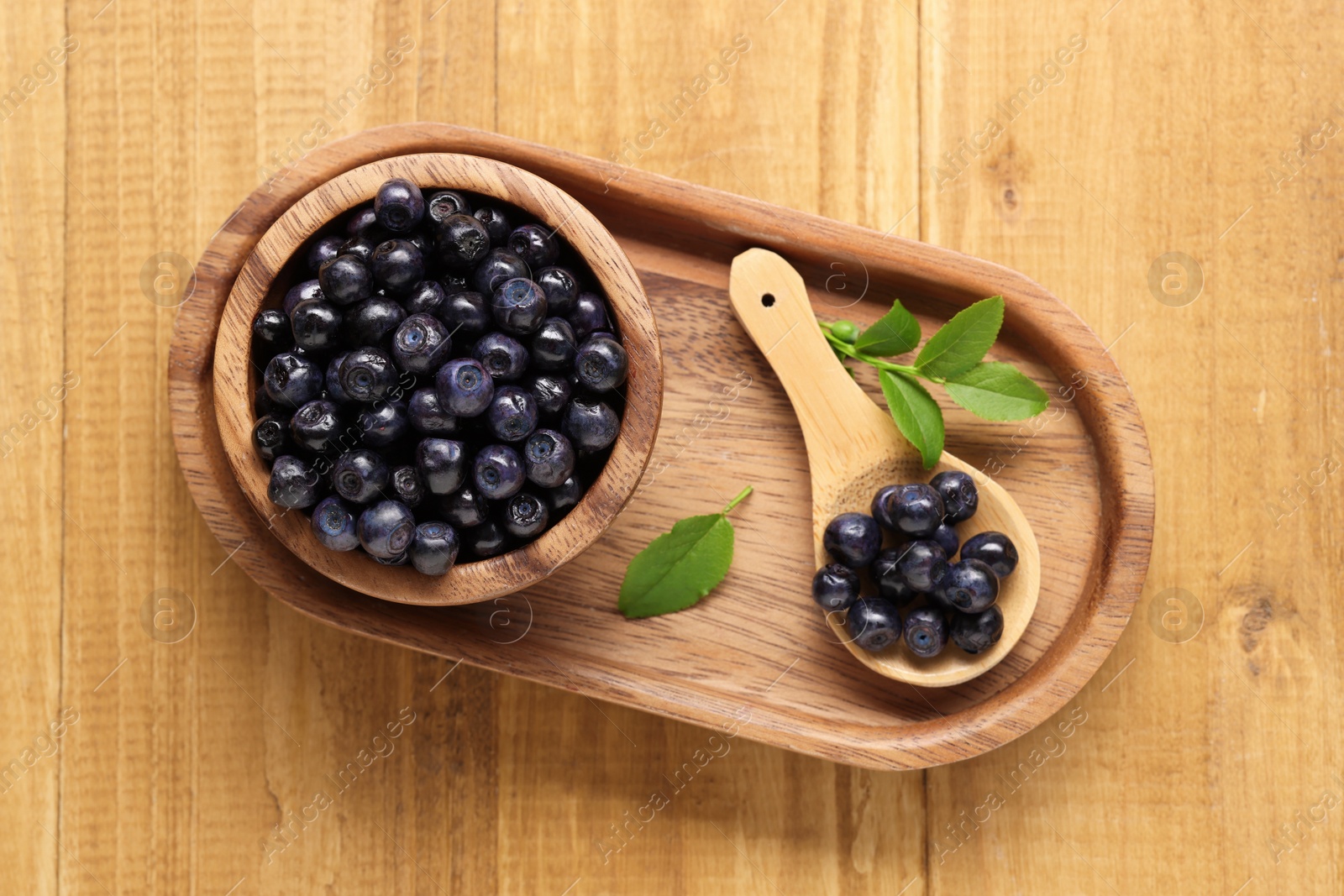 Photo of Bowl, spoon with tasty fresh bilberries and leaves on wooden table, top view