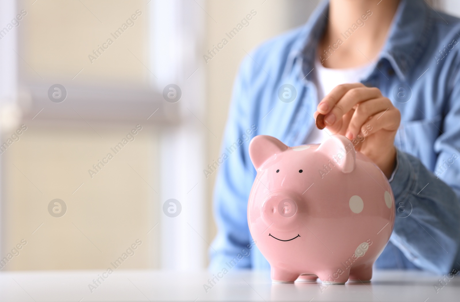 Photo of Woman putting coin into piggy bank at table indoors, closeup. Space for text