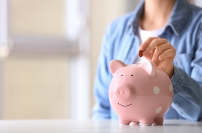 Woman putting coin into piggy bank at table indoors, closeup. Space for text