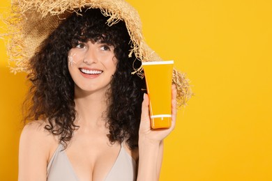 Photo of Beautiful happy woman in straw hat with sun protection cream on her face holding tube of sunscreen against orange background, space for text