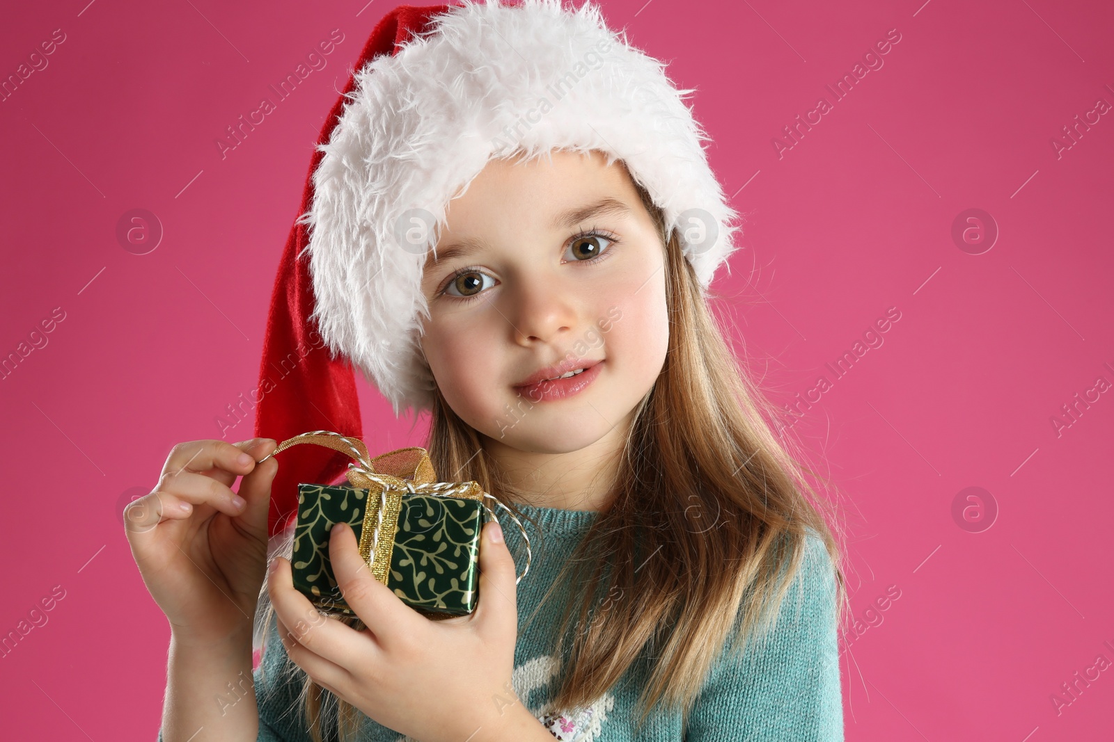 Photo of Cute child in Santa hat with Christmas gift on pink background