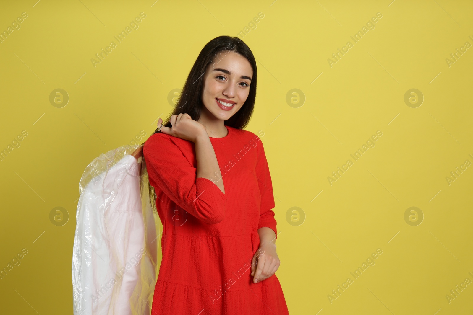 Photo of Young woman holding hanger with shirt in plastic bag on yellow background. Dry-cleaning service