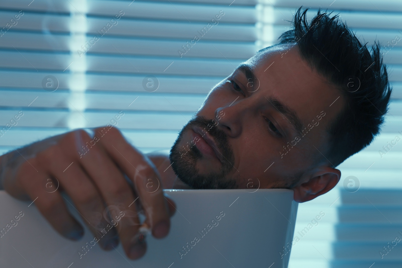 Photo of Upset man smoking in bathtub at night. Loneliness concept