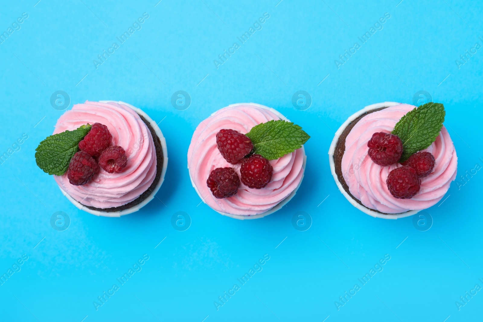 Photo of Delicious cupcakes with cream and raspberries on light blue background, flat lay