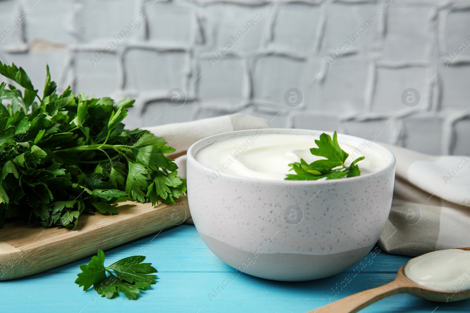 Photo of Bowl of fresh sour cream with parsley and spoon on light blue wooden table