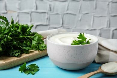 Photo of Bowl of fresh sour cream with parsley and spoon on light blue wooden table