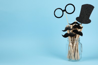 Photo of Fake paper mustaches, glasses, hat and straws in jar on light blue background. Space for text