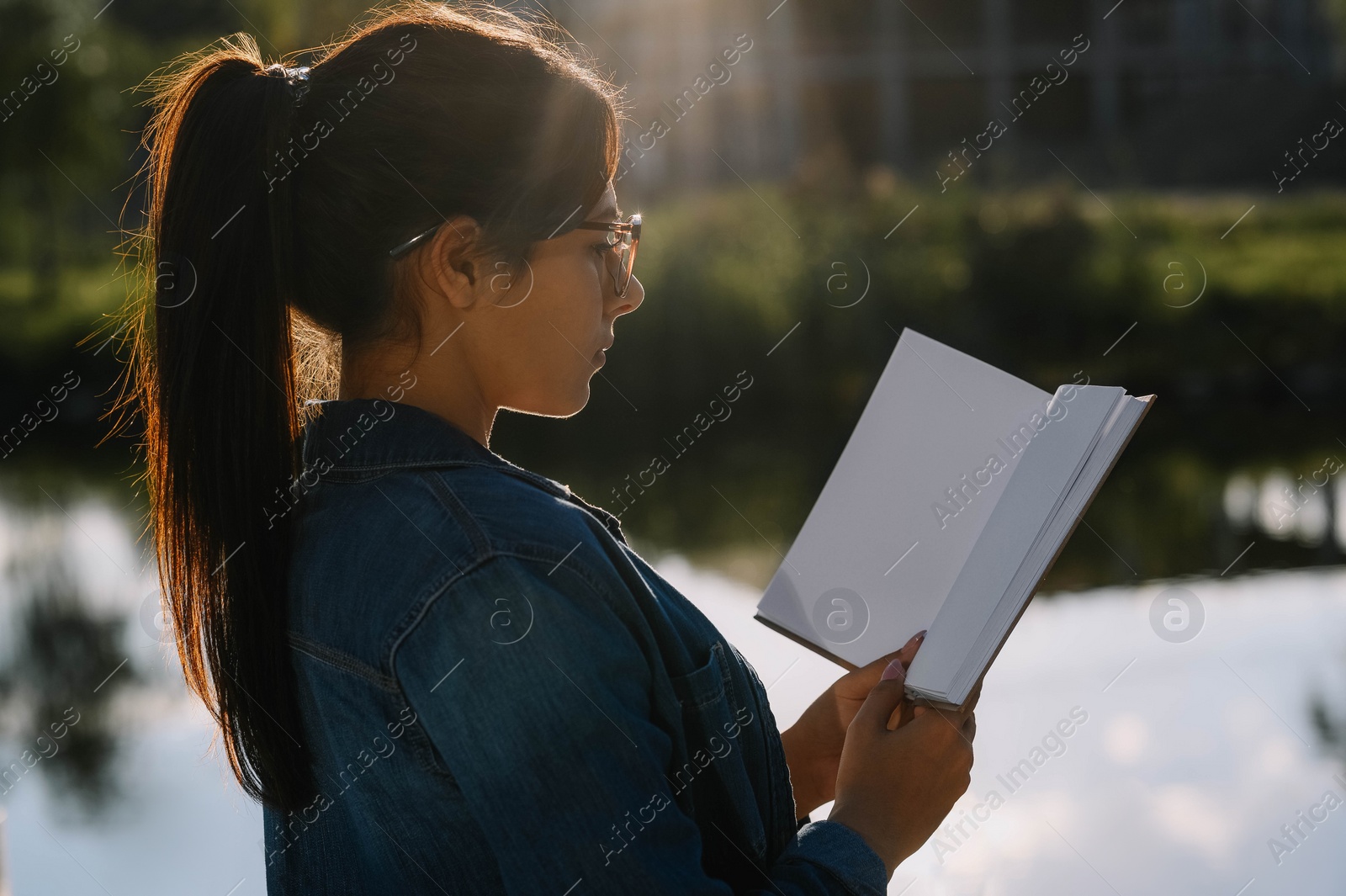 Photo of Young woman reading book near lake on sunny day