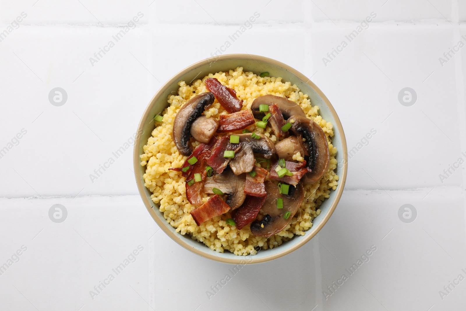 Photo of Tasty millet porridge with mushrooms, bacon and green onion in bowl on white tiled table, top view