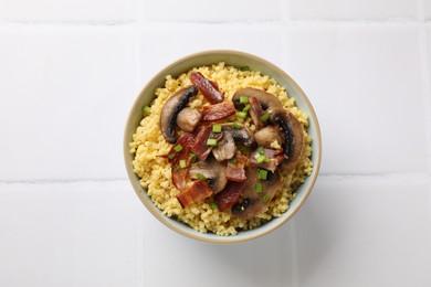 Photo of Tasty millet porridge with mushrooms, bacon and green onion in bowl on white tiled table, top view