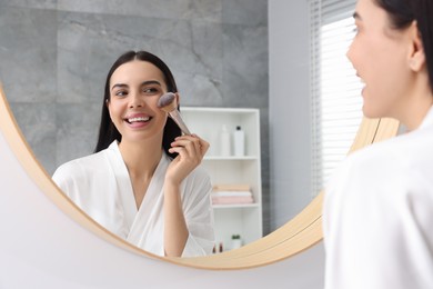 Photo of Beautiful young woman applying makeup with brush near mirror in bathroom