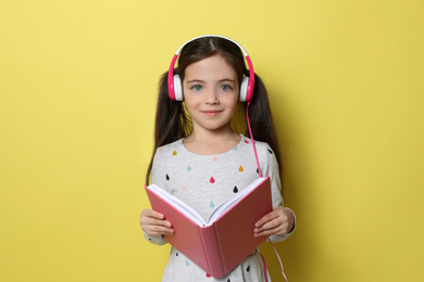 Cute little girl with headphones listening to audiobook on yellow background