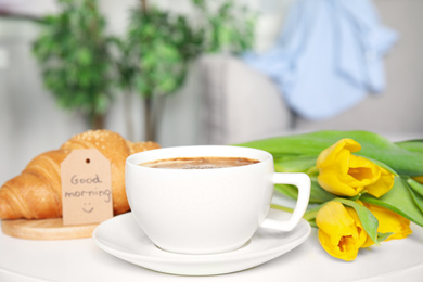 Photo of Aromatic coffee, croissant, beautiful flowers and GOOD MORNING wish on white table indoors