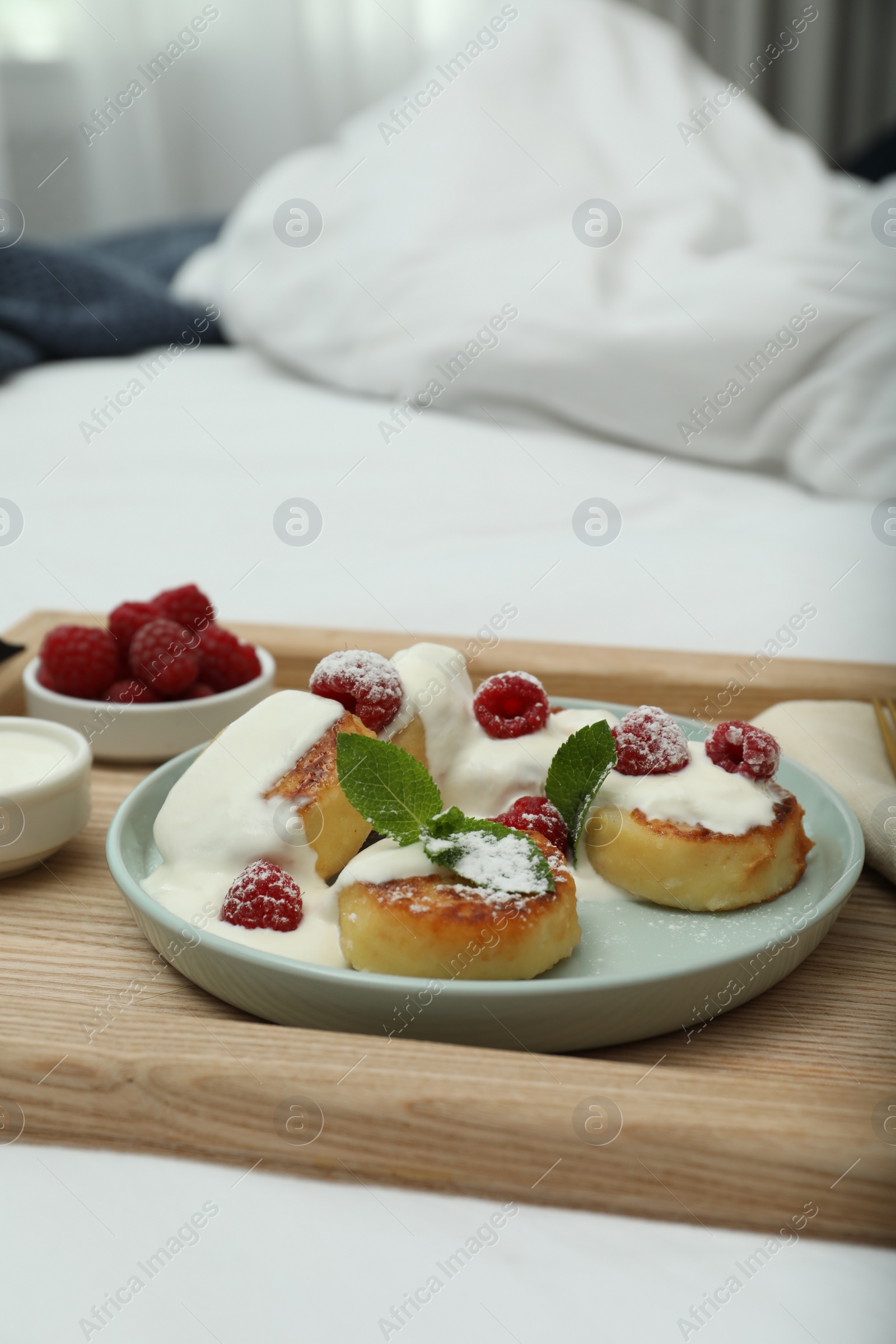 Photo of Tasty breakfast served in bedroom. Cottage cheese pancakes with fresh raspberries, mint, sour cream and icing sugar on wooden tray