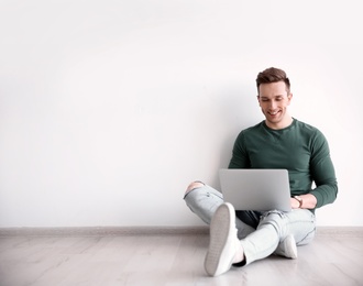 Young blogger with laptop sitting on floor against light wall