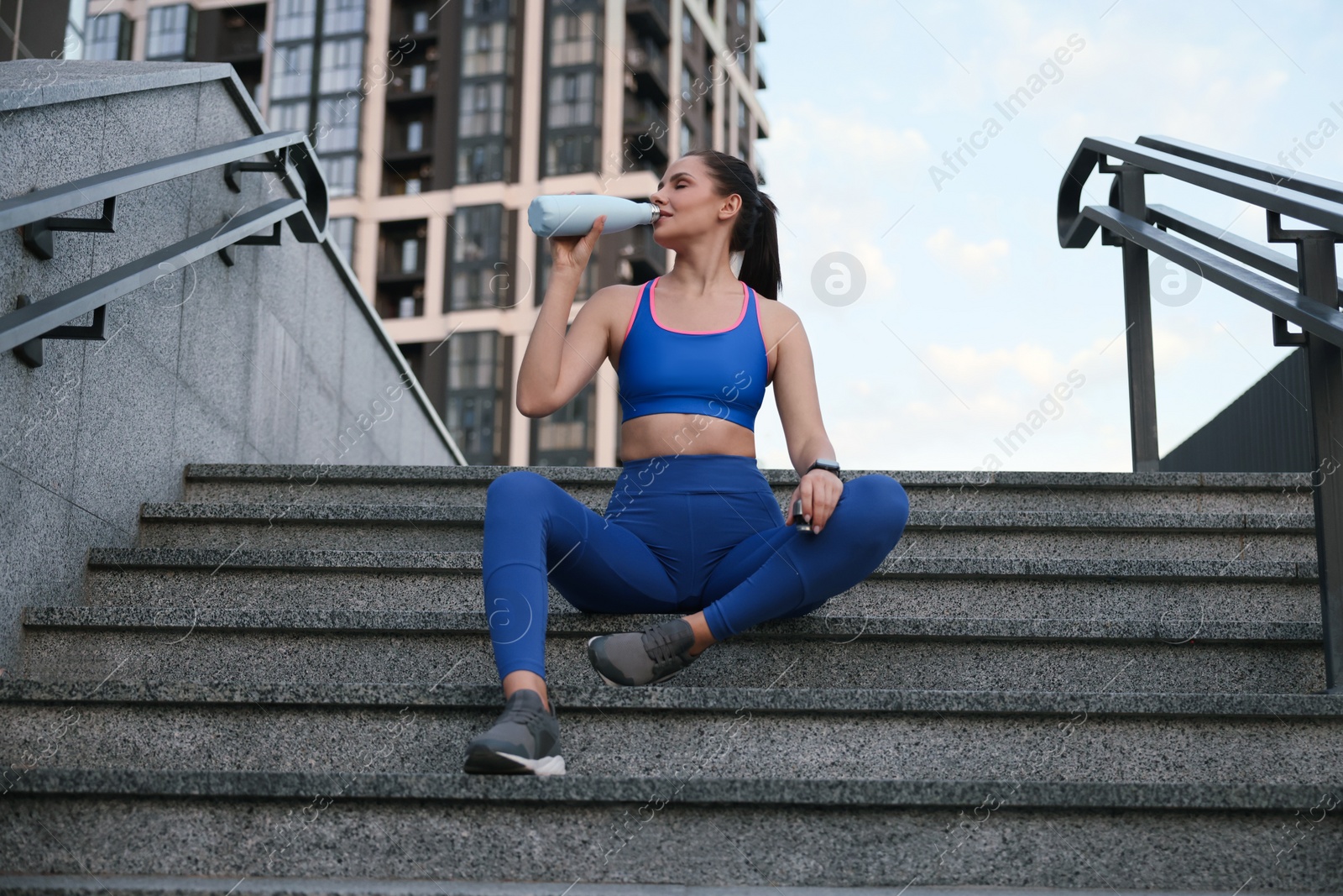 Photo of Beautiful woman in stylish sportswear drinking water on stairs outdoors