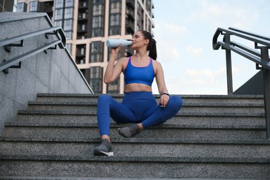 Beautiful woman in stylish sportswear drinking water on stairs outdoors