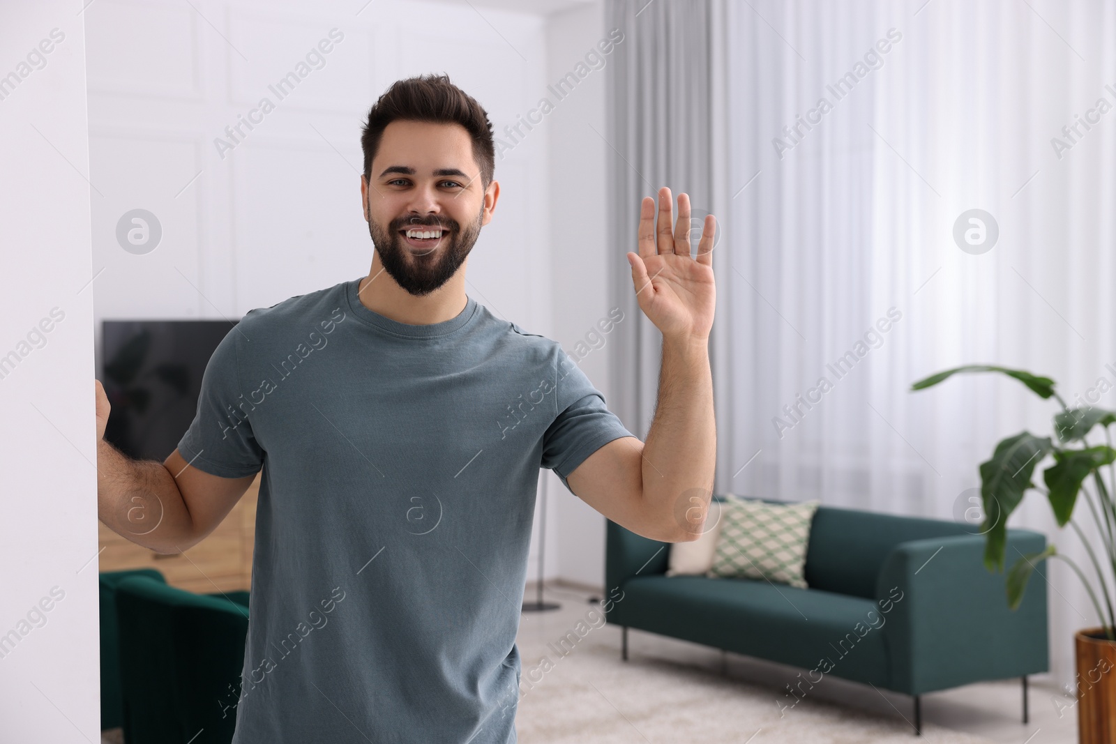 Photo of Happy man waving near white wall at home, space for text. Invitation to come in room