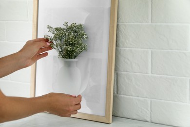 Woman attaching silicone vase with flowers to picture frame's glass on white table, closeup. Space for text