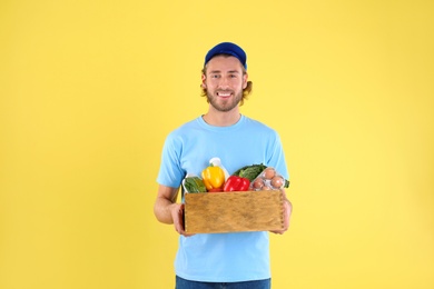 Delivery man holding wooden crate with food products on color background