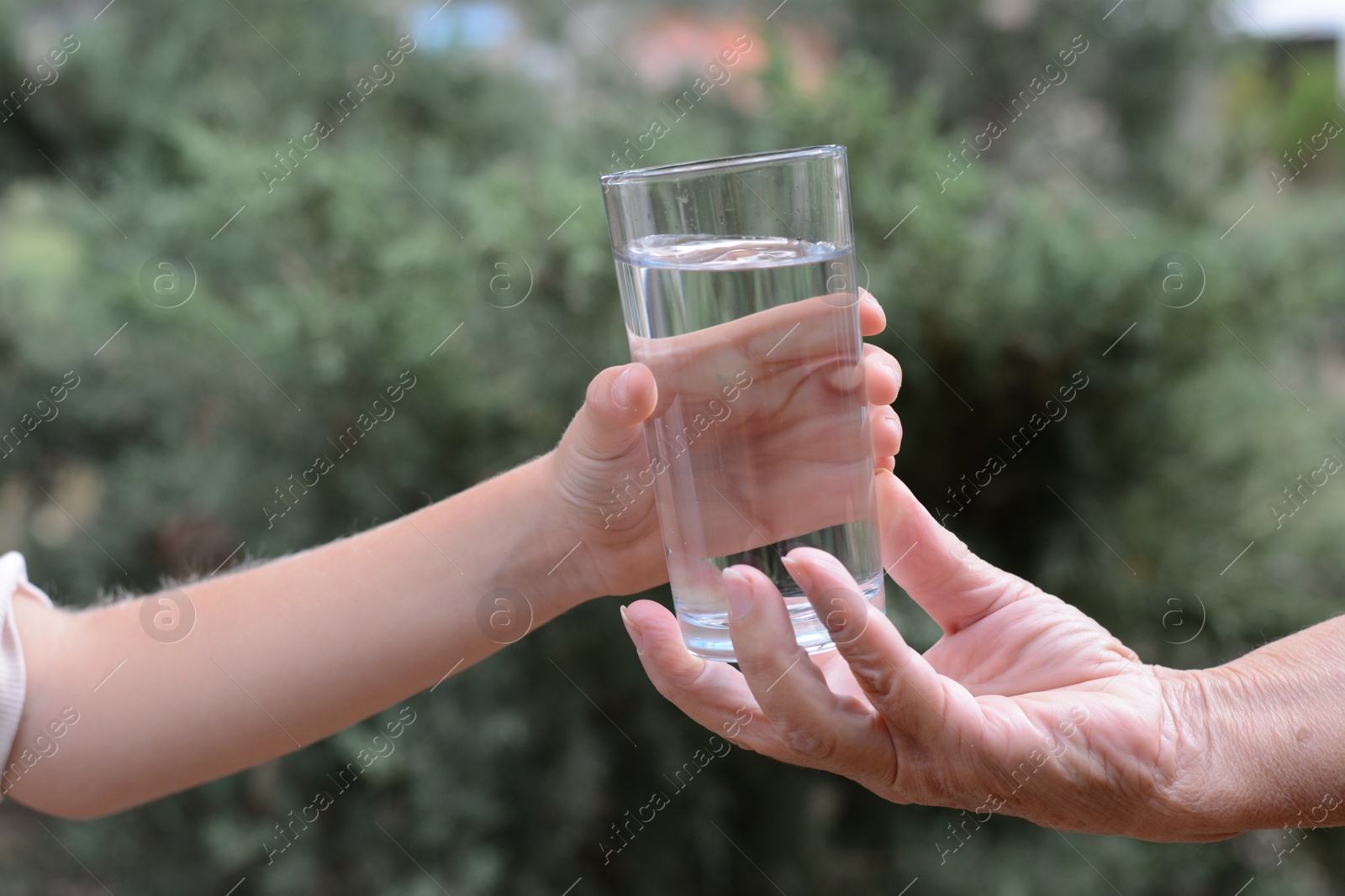 Photo of Child giving glass of water to elderly woman outdoors, closeup