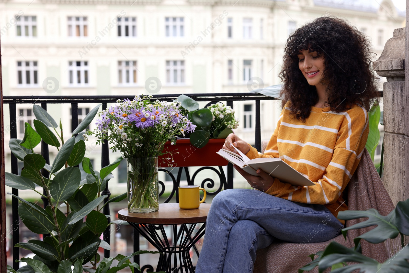 Photo of Young woman reading book at table on balcony with beautiful houseplants