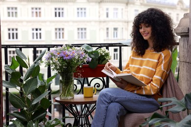 Photo of Young woman reading book at table on balcony with beautiful houseplants