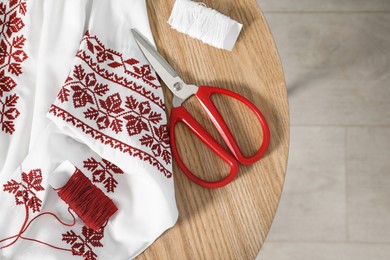 Photo of White shirt with beautiful embroidery design, threads and scissors on wooden table, flat lay. Ukrainian national clothes