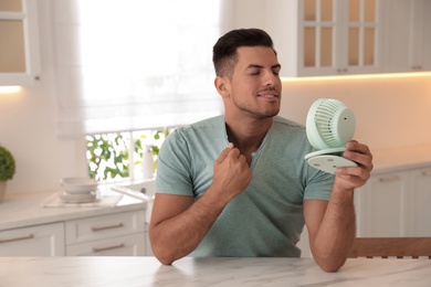 Photo of Man enjoying air flow from portable fan at table in kitchen. Summer heat