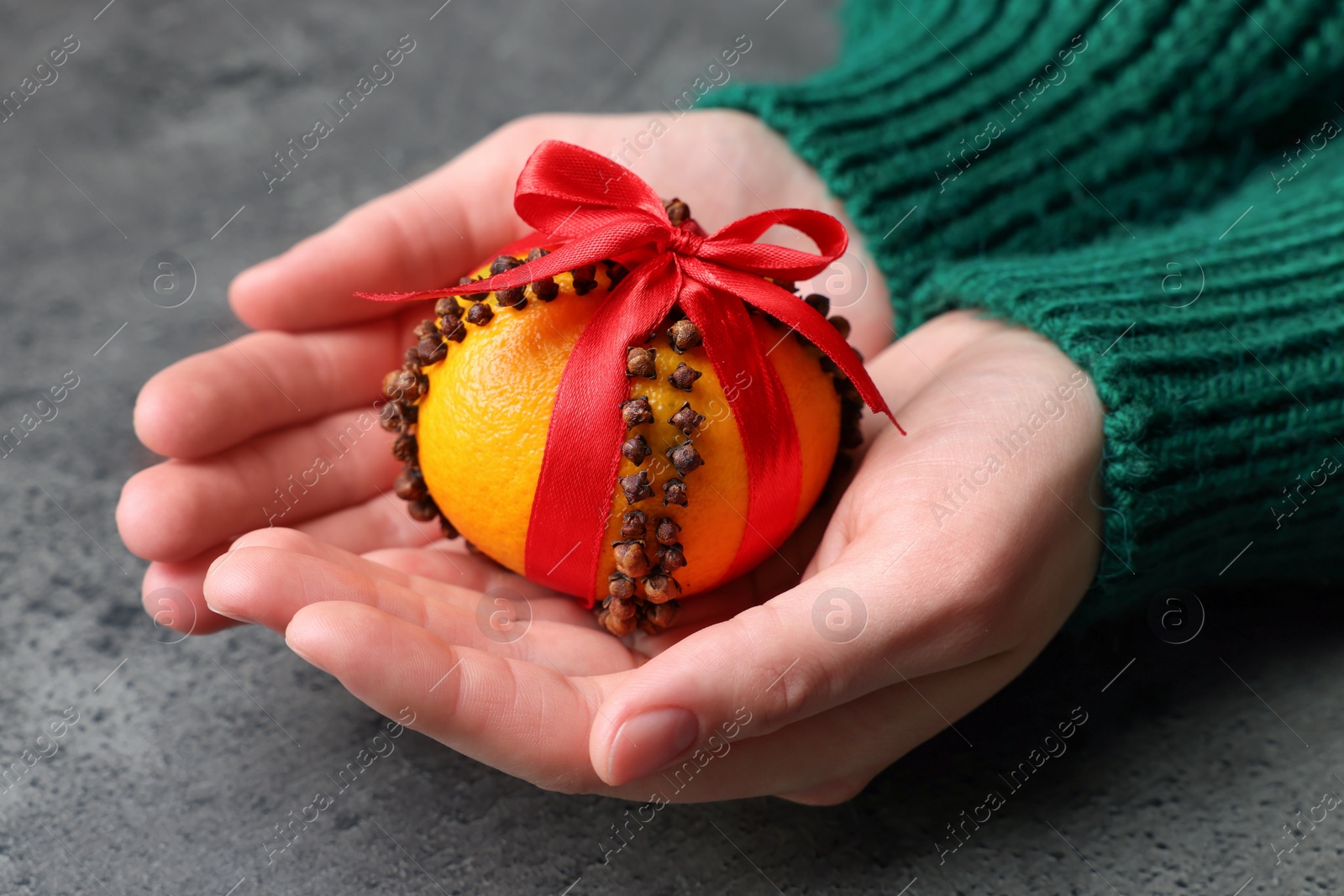 Photo of Woman holding pomander ball with red ribbon made of fresh tangerine and cloves at grey table, closeup
