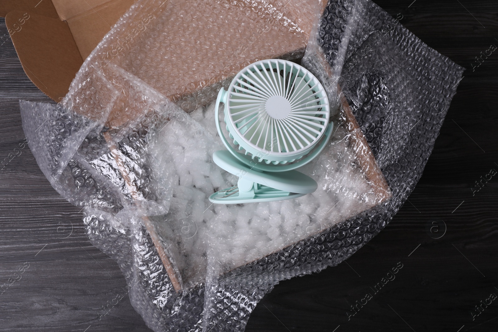 Photo of Small fan with bubble wrap and packaging foam in cardboard box on dark wooden table, top view