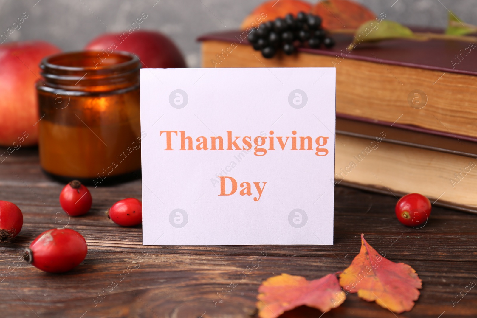 Photo of Paper card with words Thanksgiving Day near autumn leaves, berries, books and candle on wooden table, closeup