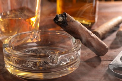Photo of Smoldering cigar, ashtray and whiskey on wooden table, closeup